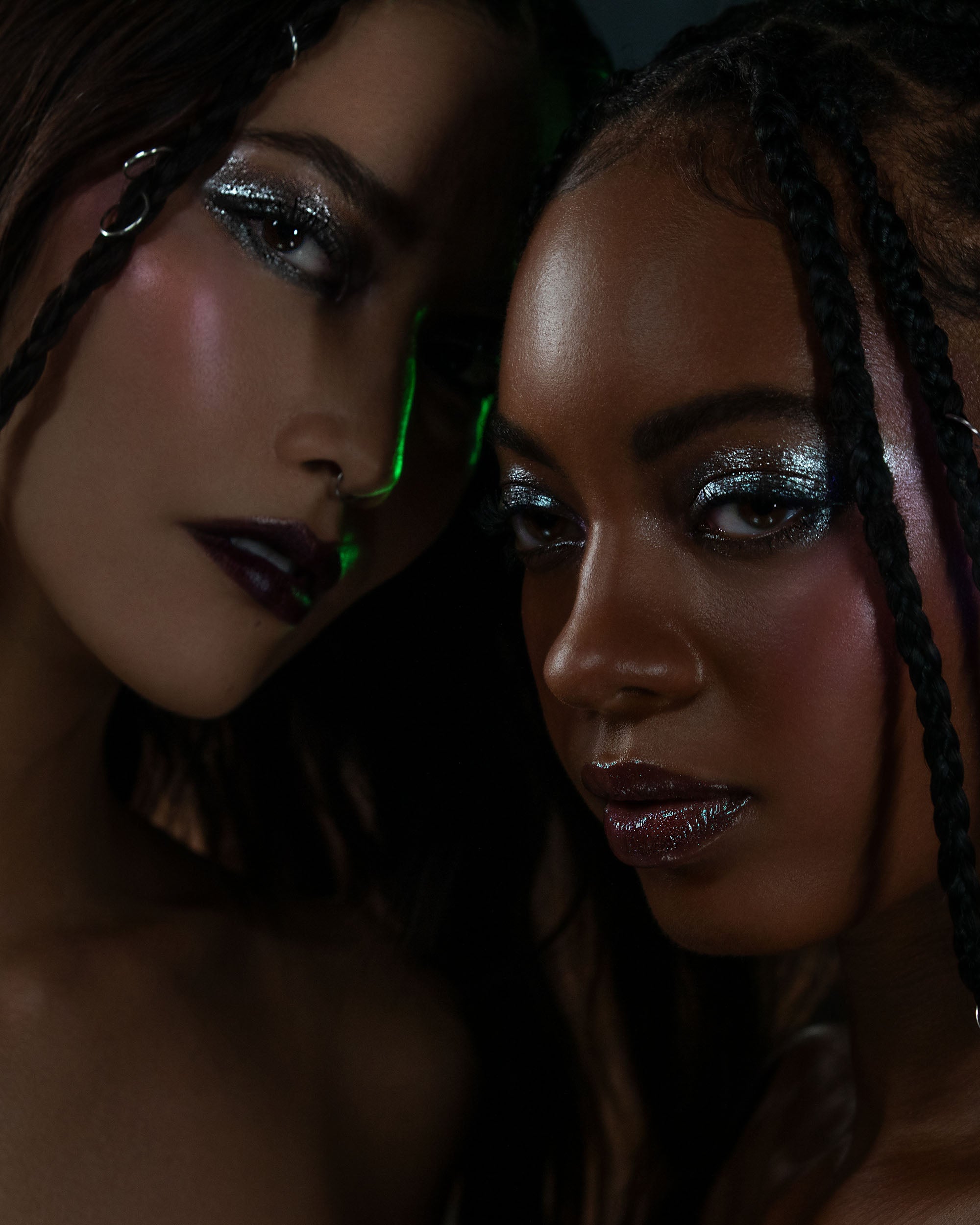 Two women with glittery makeup and braided hair in a dark setting.