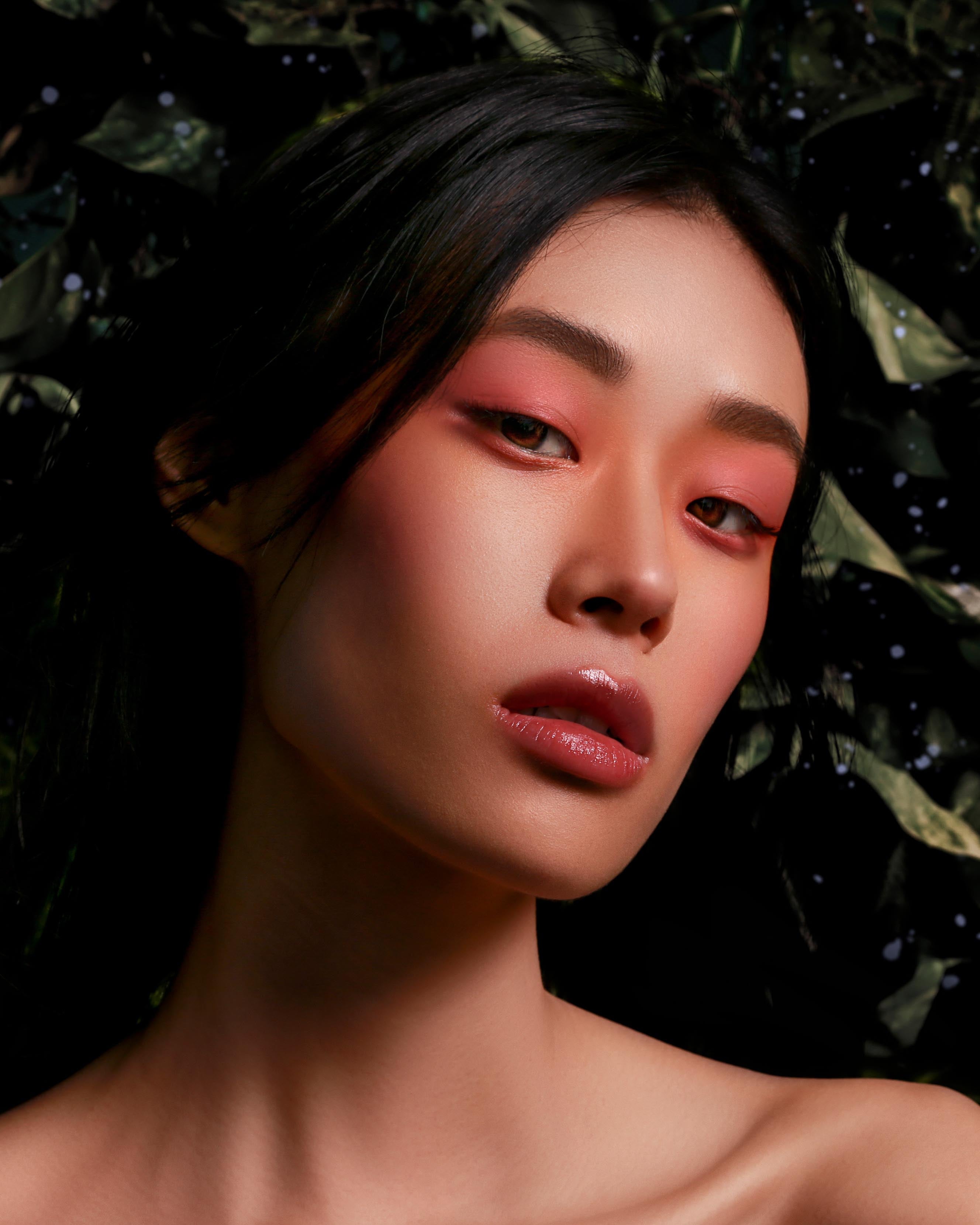 Model with light skin wearing pink eye, lip, and cheek makeup against a dark leafy background