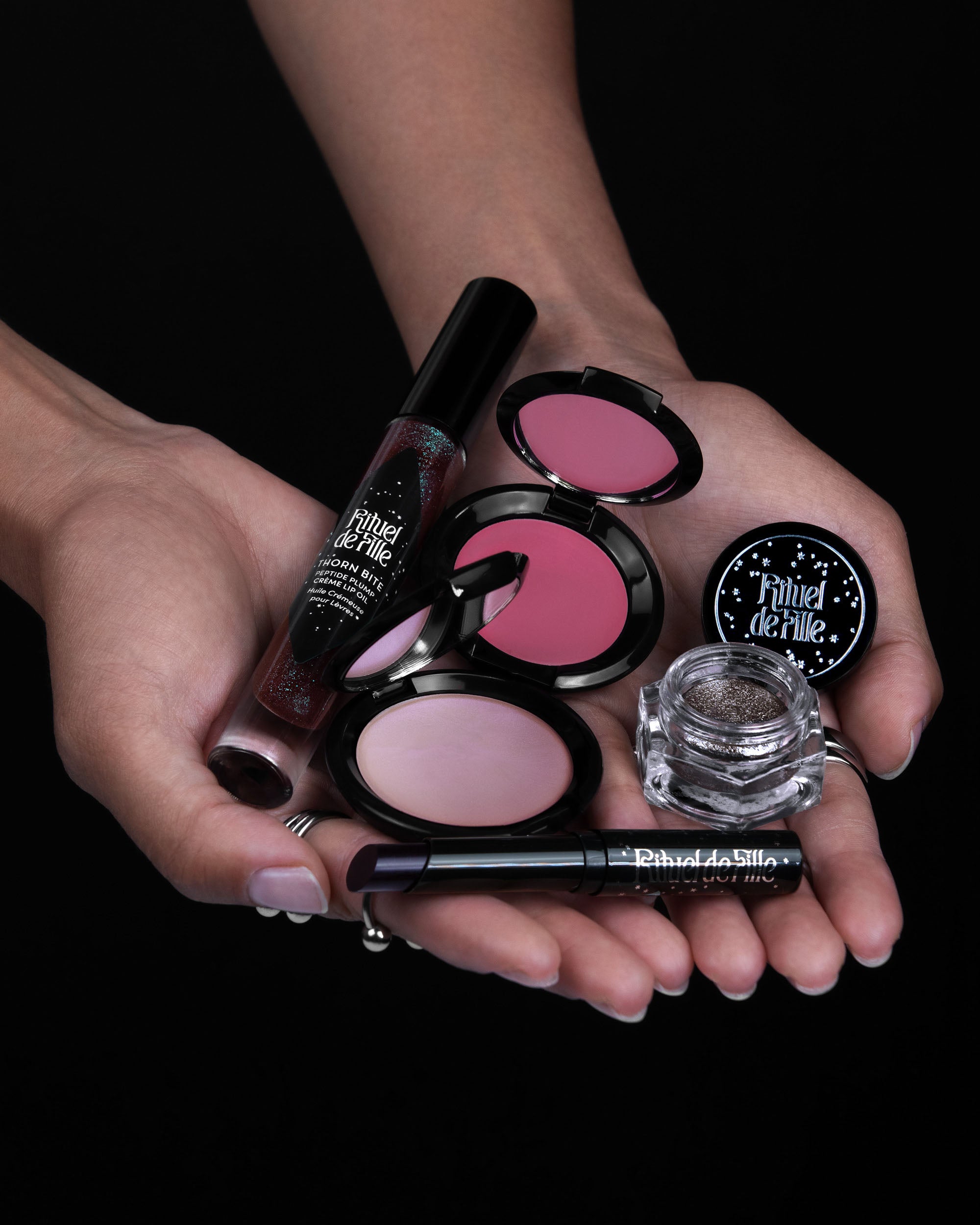 Makeup products including blushes, eyeliner, and mascara held in hands against a black background.