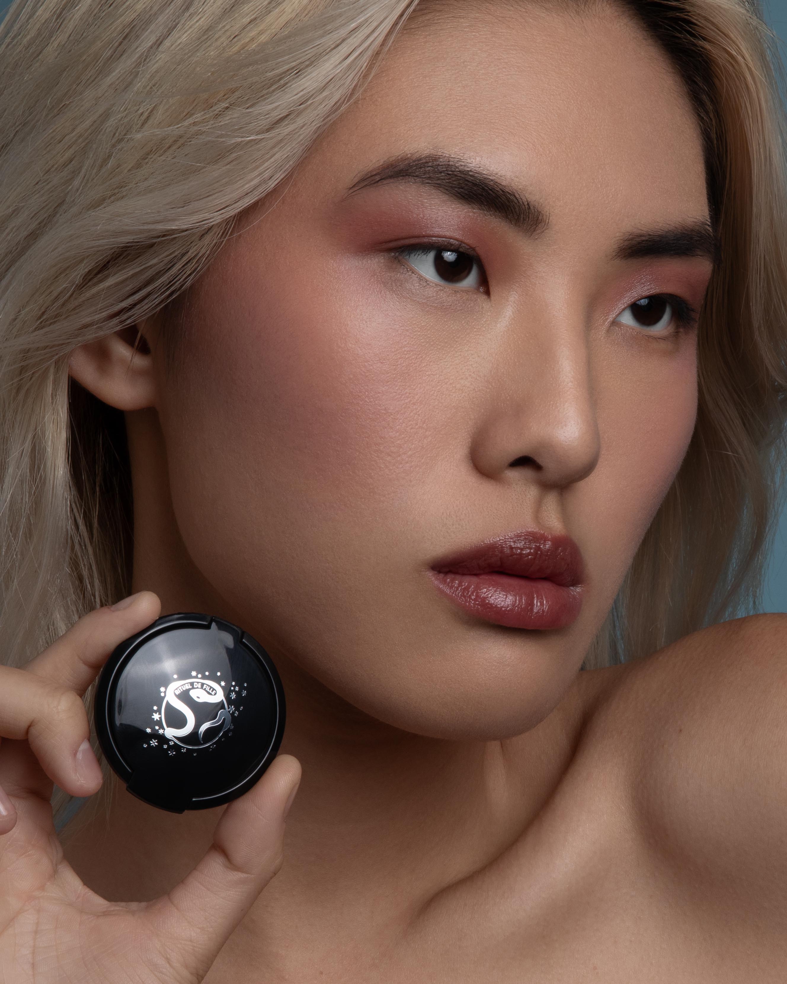 Close-up of a model with medium skin wearing rose pink makeup on lips and cheeks holding a compact