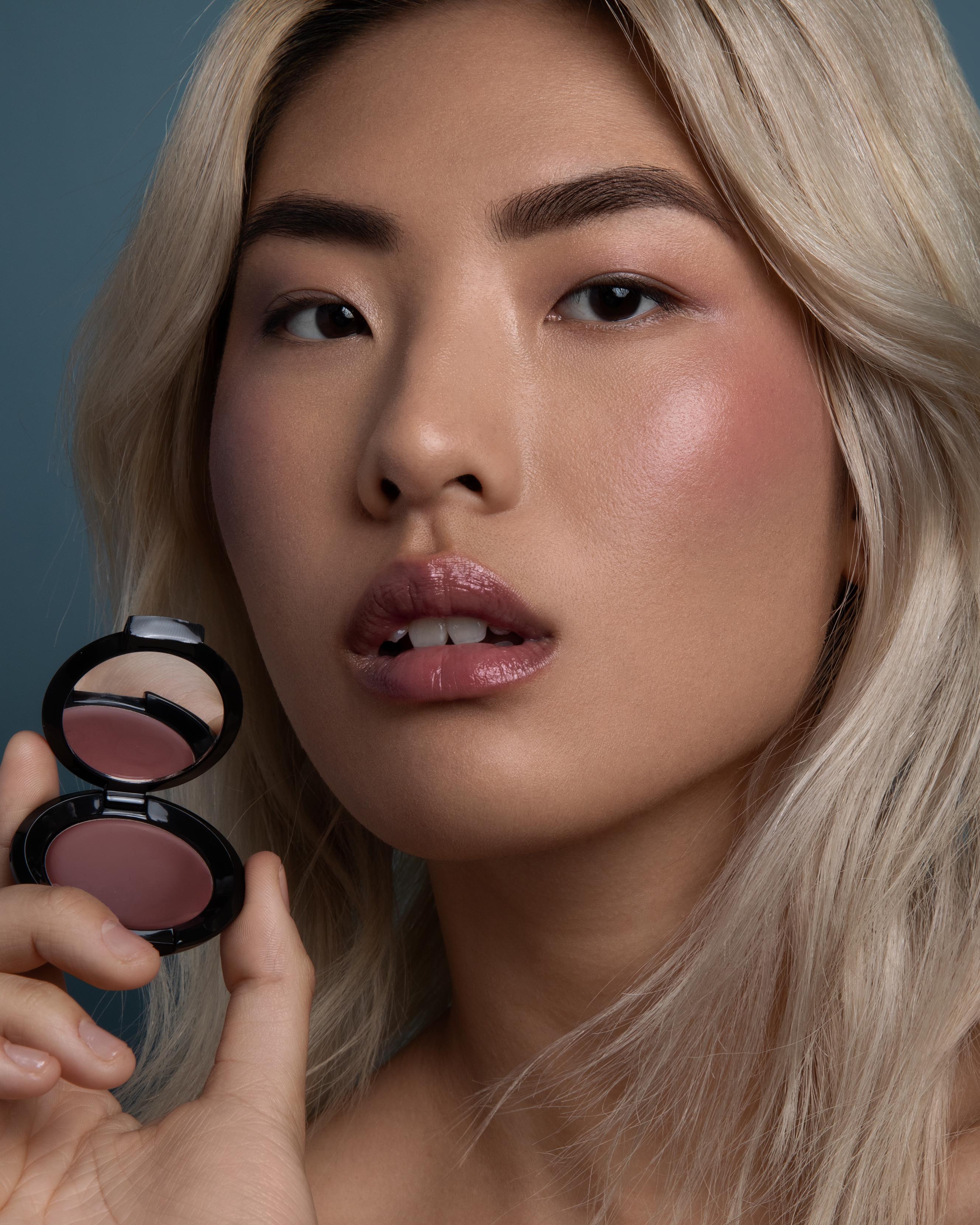 Close-up of a model with medium skin holding a compact and wearing nude pink makeup on lips, eyes, and cheeks