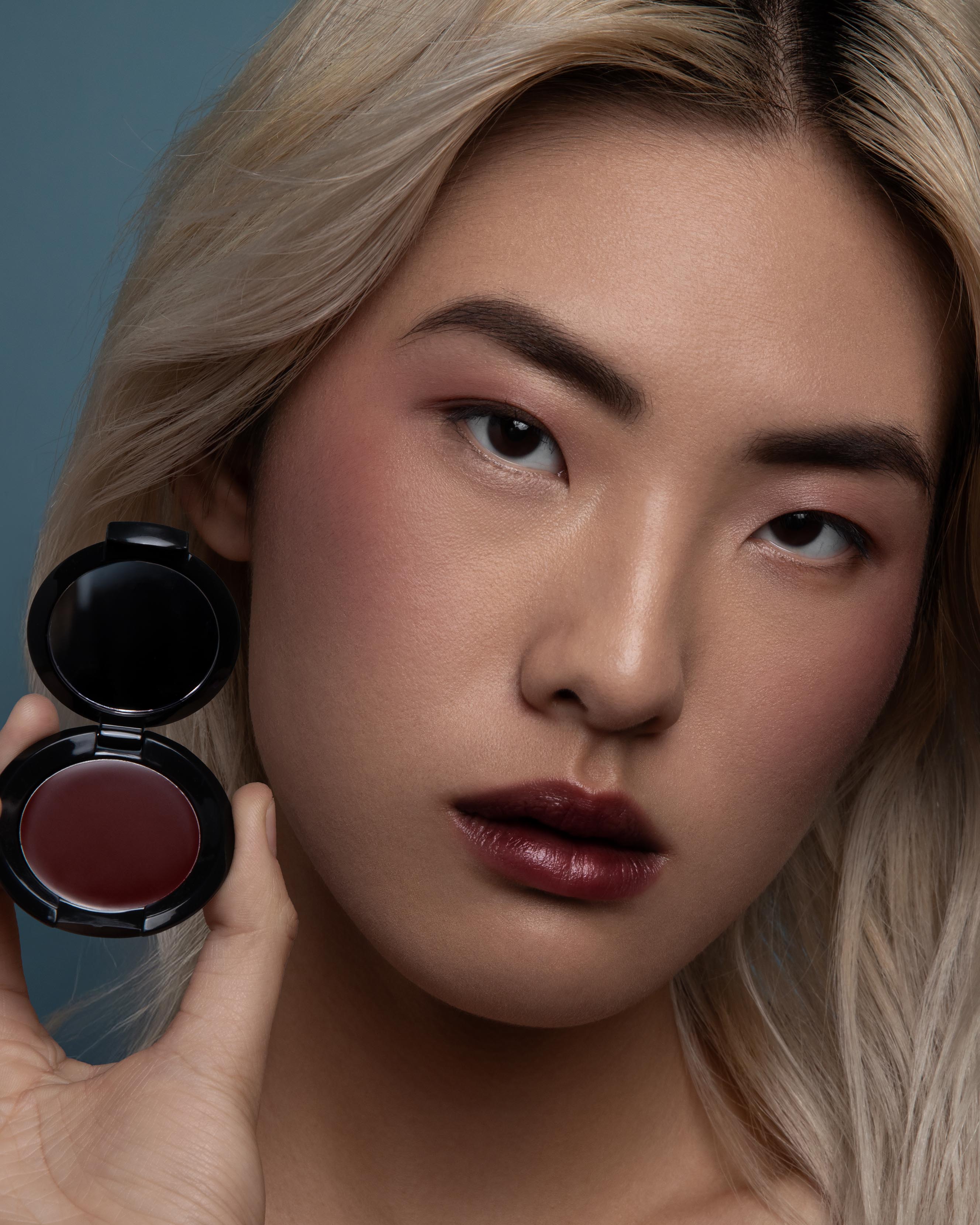 Close-up of a model with medium skin holding a compact and wearing burgundy red makeup on lips, eyes, and cheeks